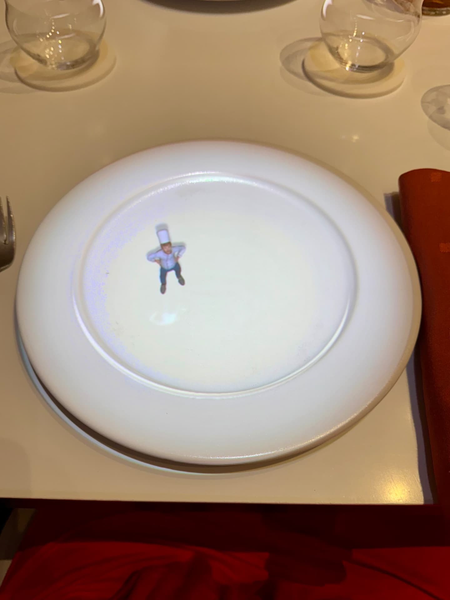 Fine dining plate with small illustrated chef figure in center, formal place setting with glasses and napkin.