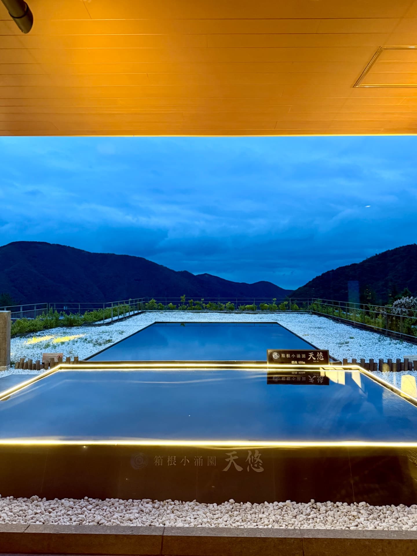 Indoor hot spring bath with mountain valley view at dusk, Japanese onsen resort with illuminated pool overlooking landscape.