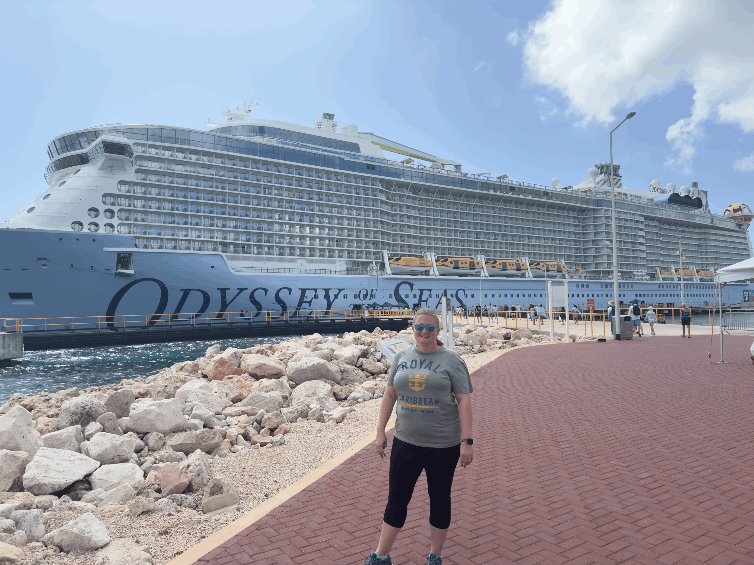 Person posing in front of Odyssey of the Seas cruise ship docked at port with rocky shoreline and clear blue sky.