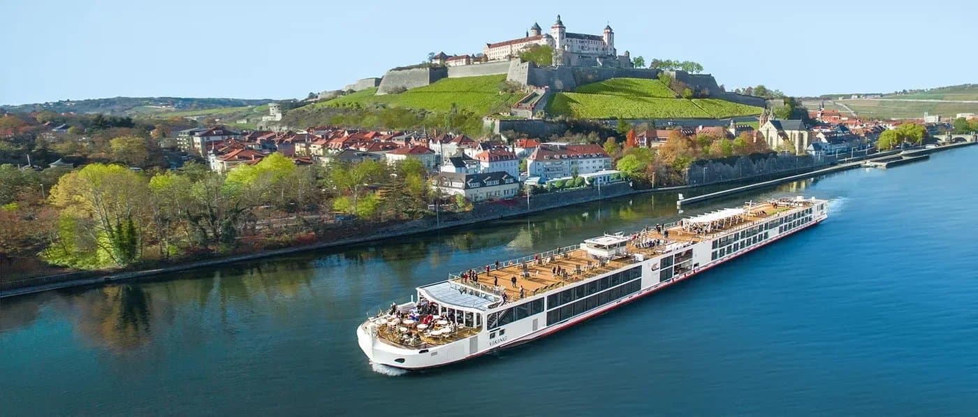 River cruise ship on blue water with medieval castle on hilltop overlooking historic German riverside town with red-roofed buildings.