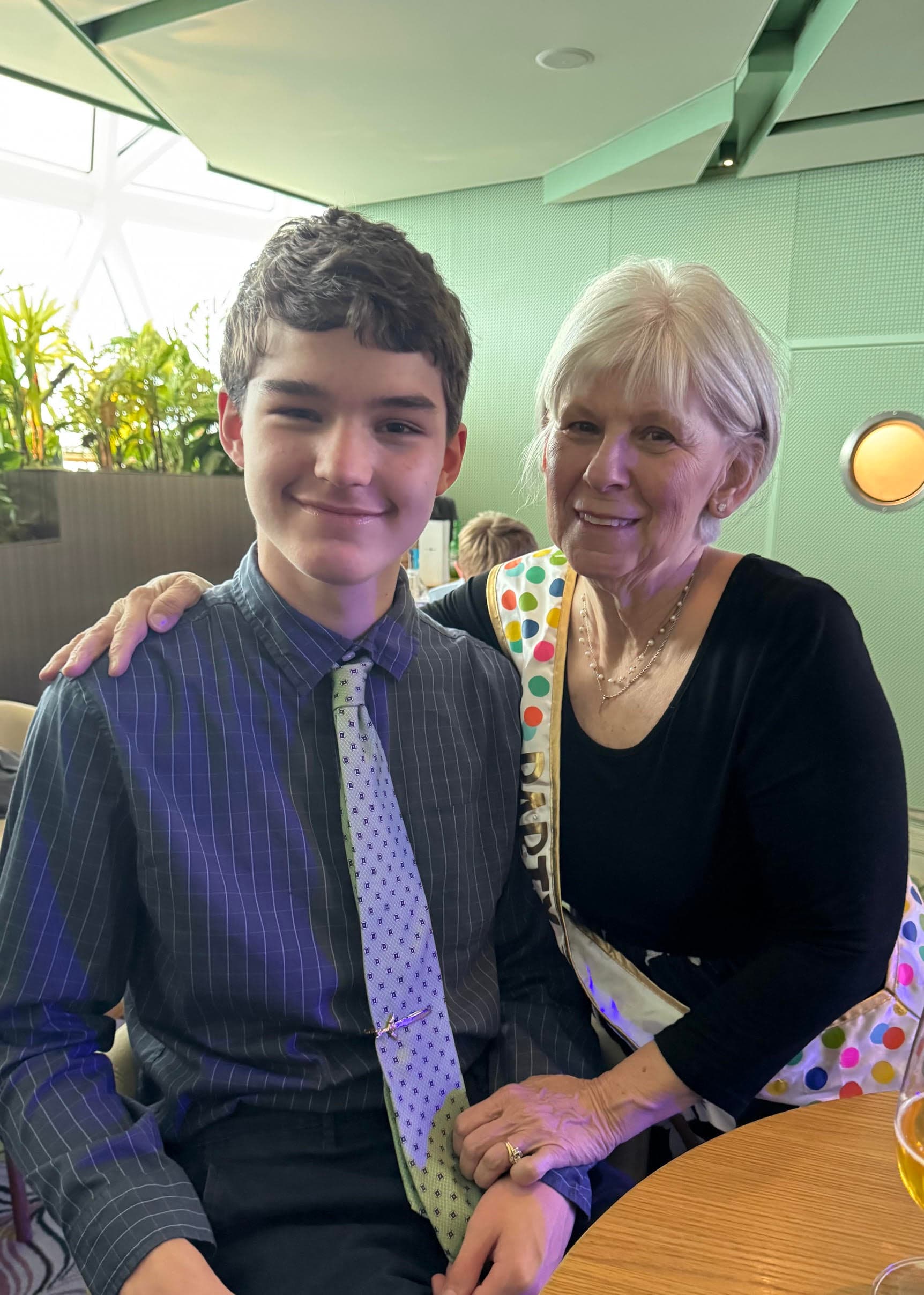 A young boy in a pinstriped suit and tie poses with an older woman in a black top at a cruise ship event.