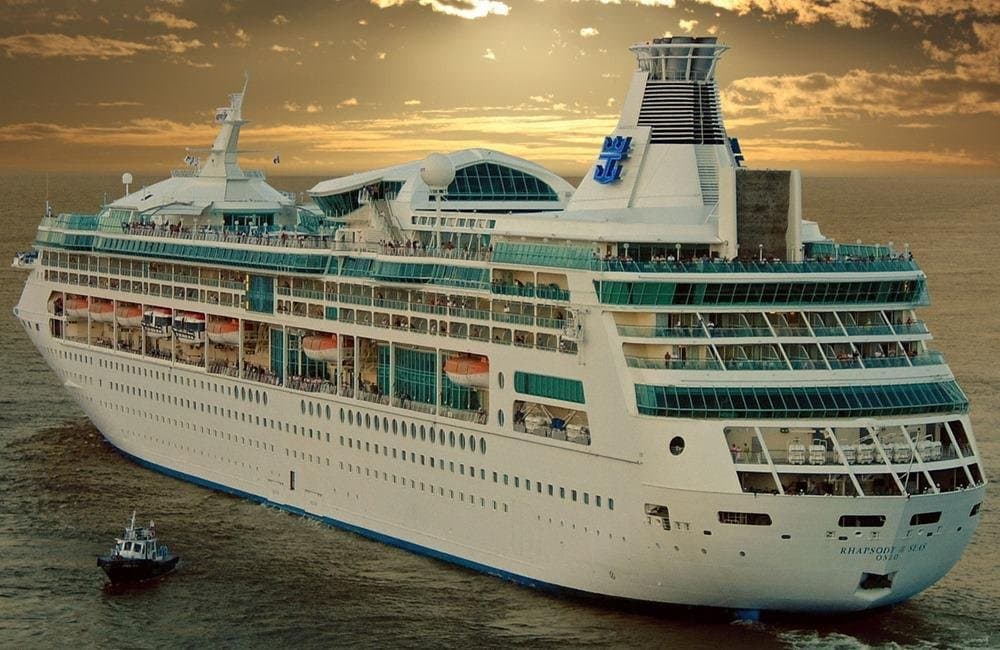 Large white and turquoise cruise ship at sea during golden hour sunset with smaller tugboat nearby.