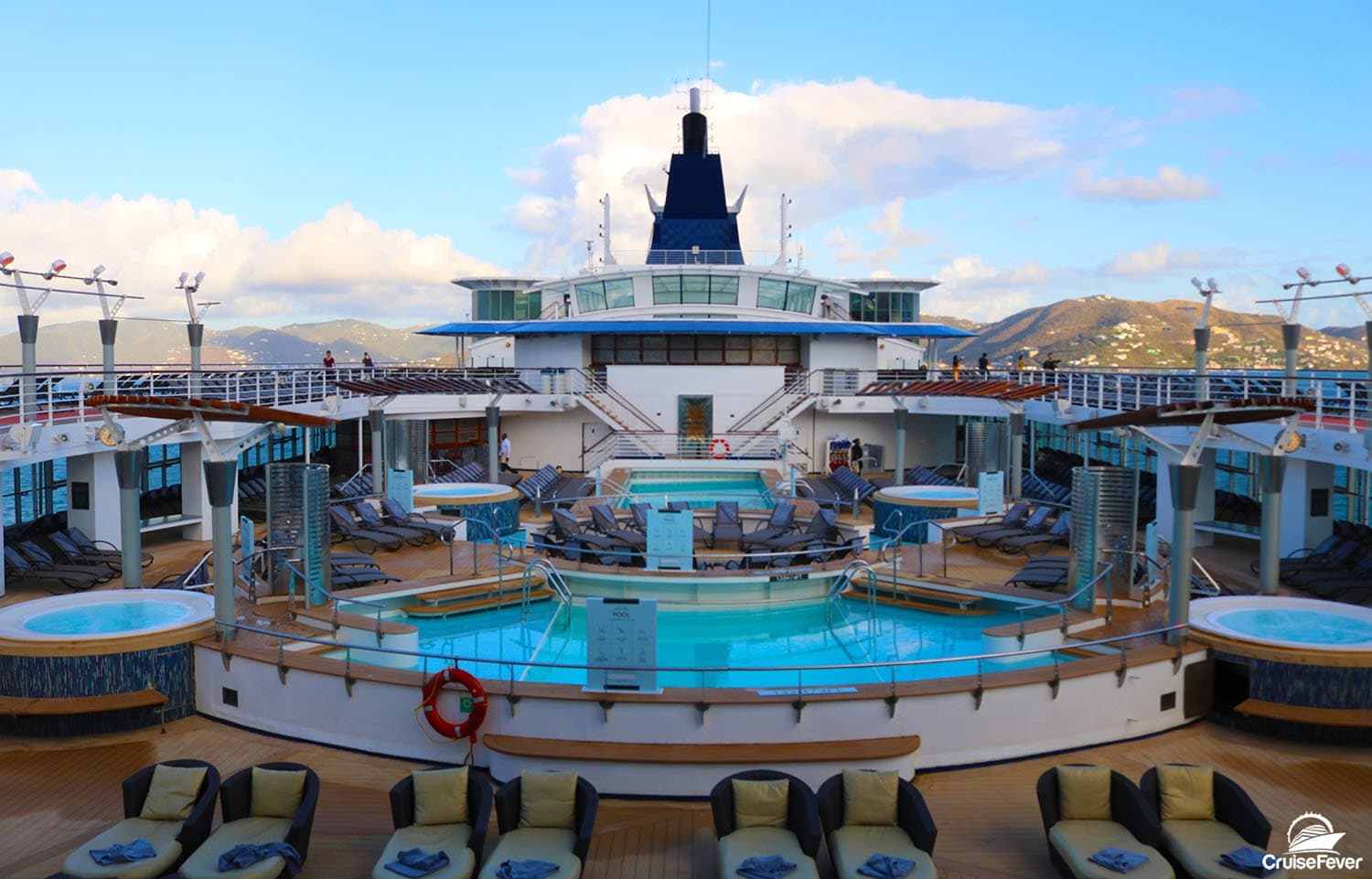 Cruise ship main pool deck with loungers, hot tubs, and mountainous coastal landscape in background