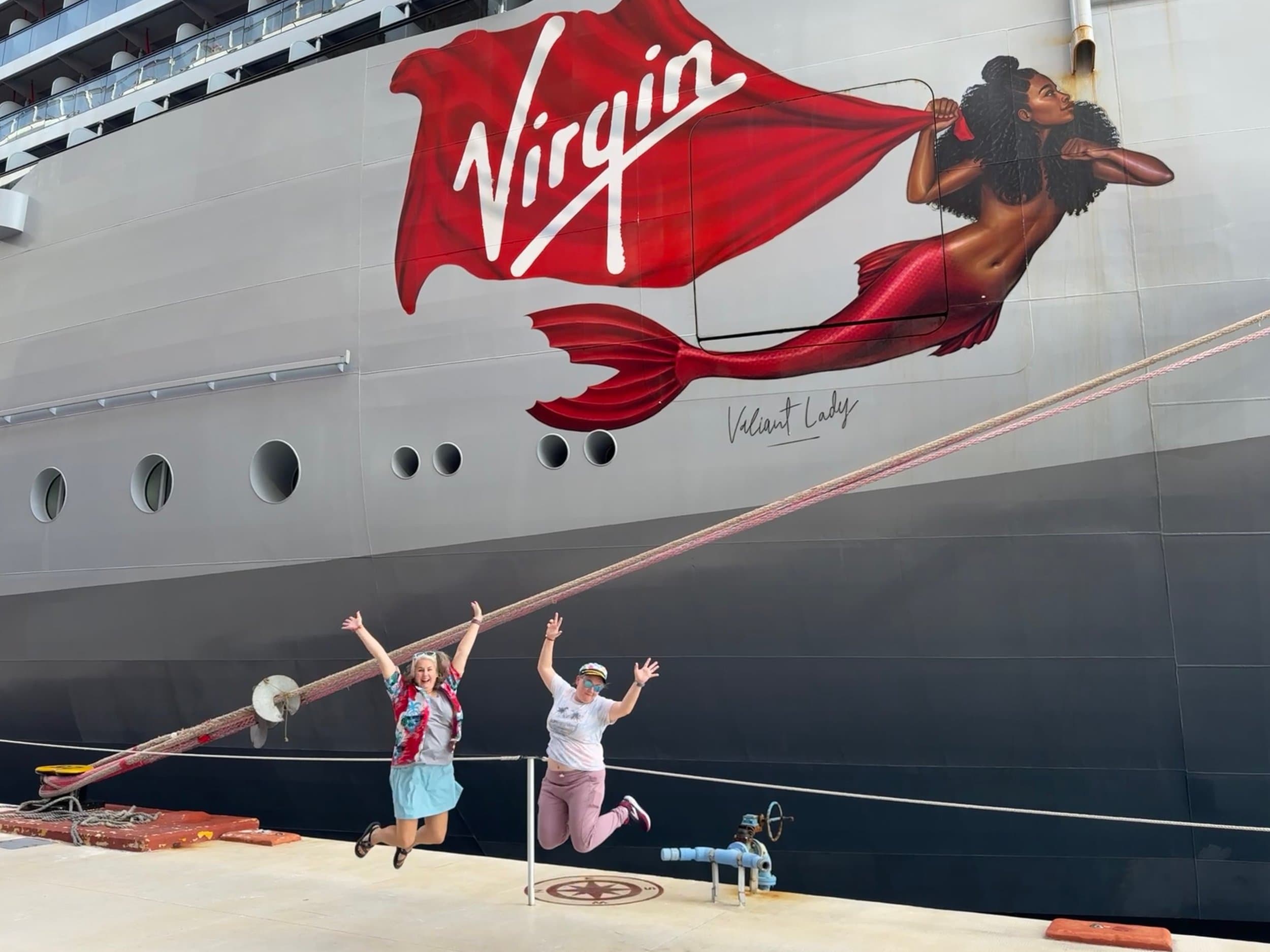 Two women posing joyfully in front of Virgin Voyages cruise ship with mermaid logo and "Valiant Lady" hull art.