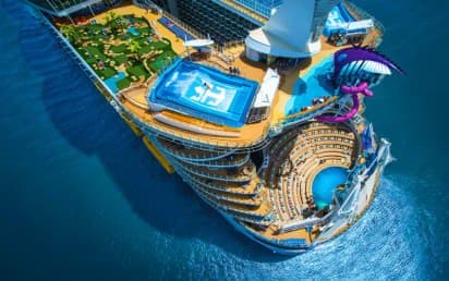 Aerial view of a large cruise ship with blue pools, water slides, and golden hull design in turquoise ocean waters.