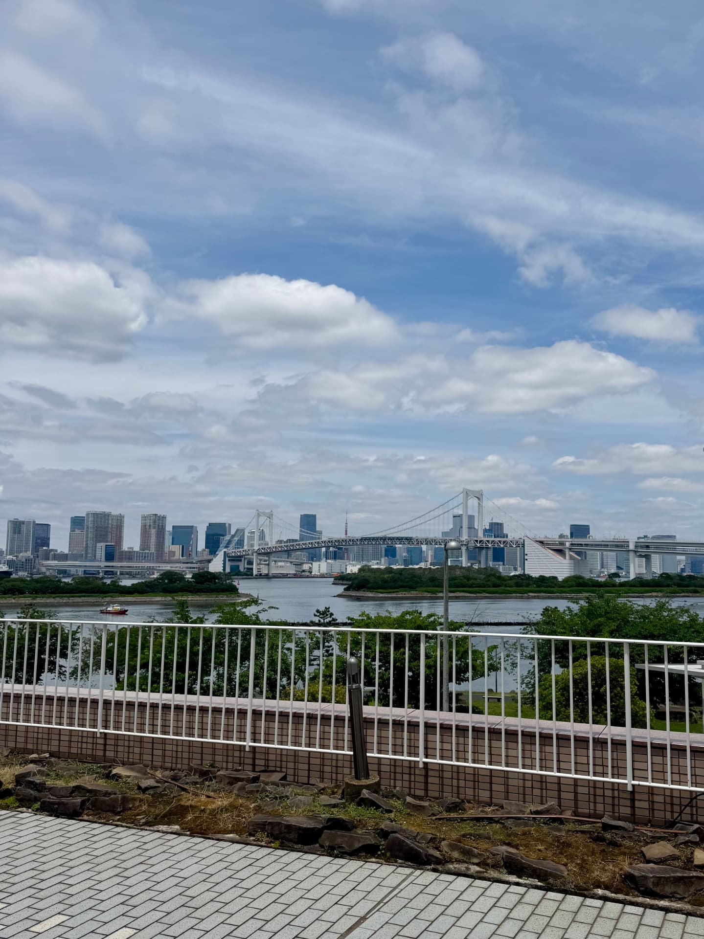 View of Tokyo skyline and Rainbow Bridge from waterfront promenade with white railings and green vegetation.
