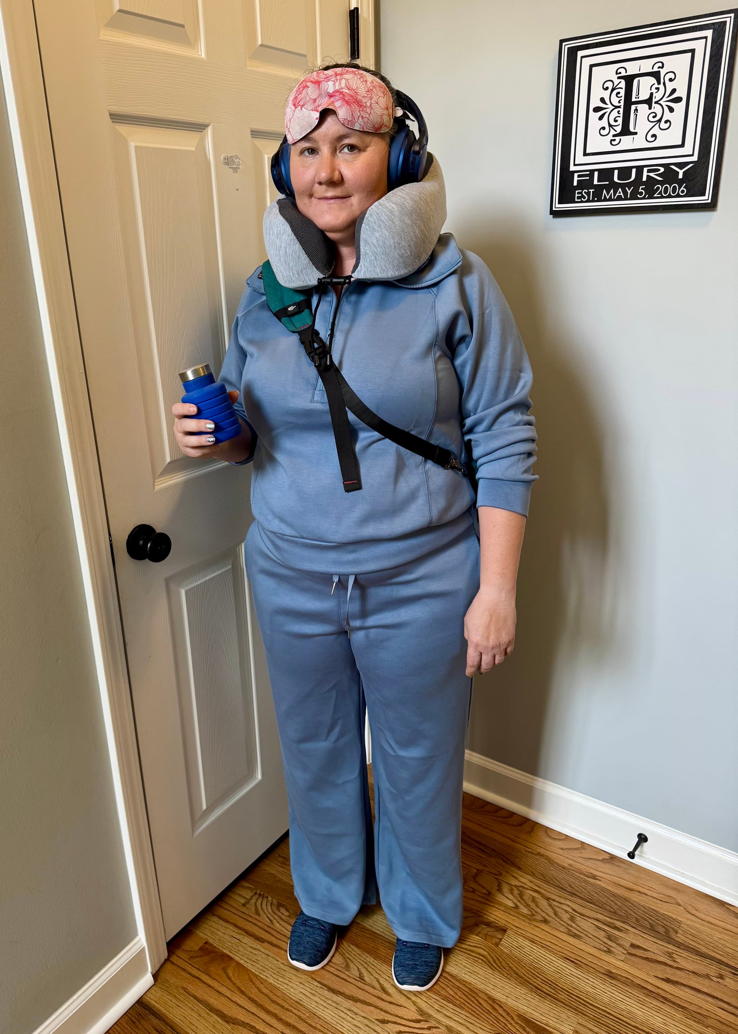 Woman in blue cruise ship crew uniform wearing headphones, neck pillow, and sleep mask, holding a beverage in an indoor hallway.