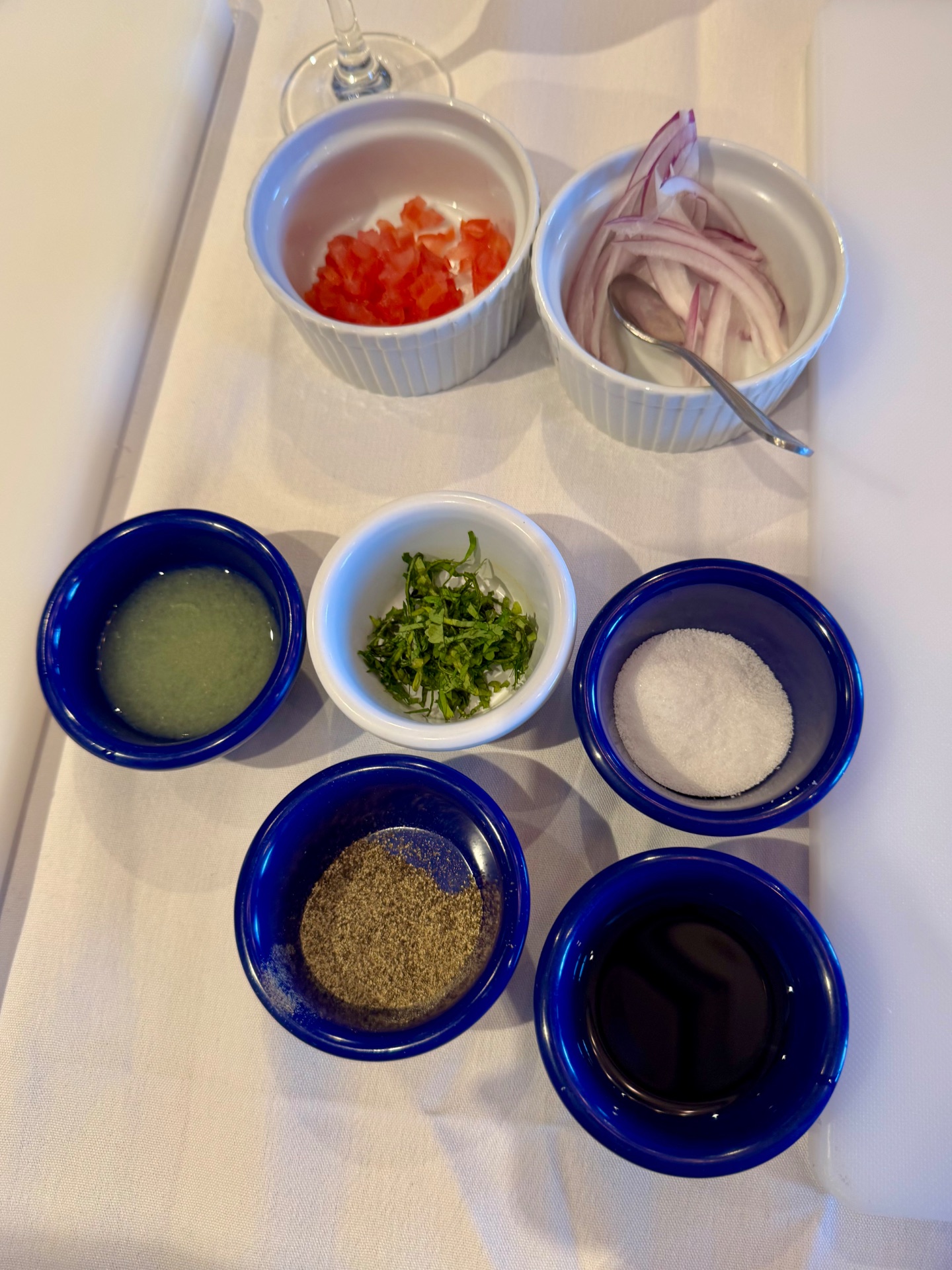 Mise en place ingredients in small bowls including diced red peppers, sliced onions, herbs, oil, salt, pepper, and dark sauce on a white tablecloth.