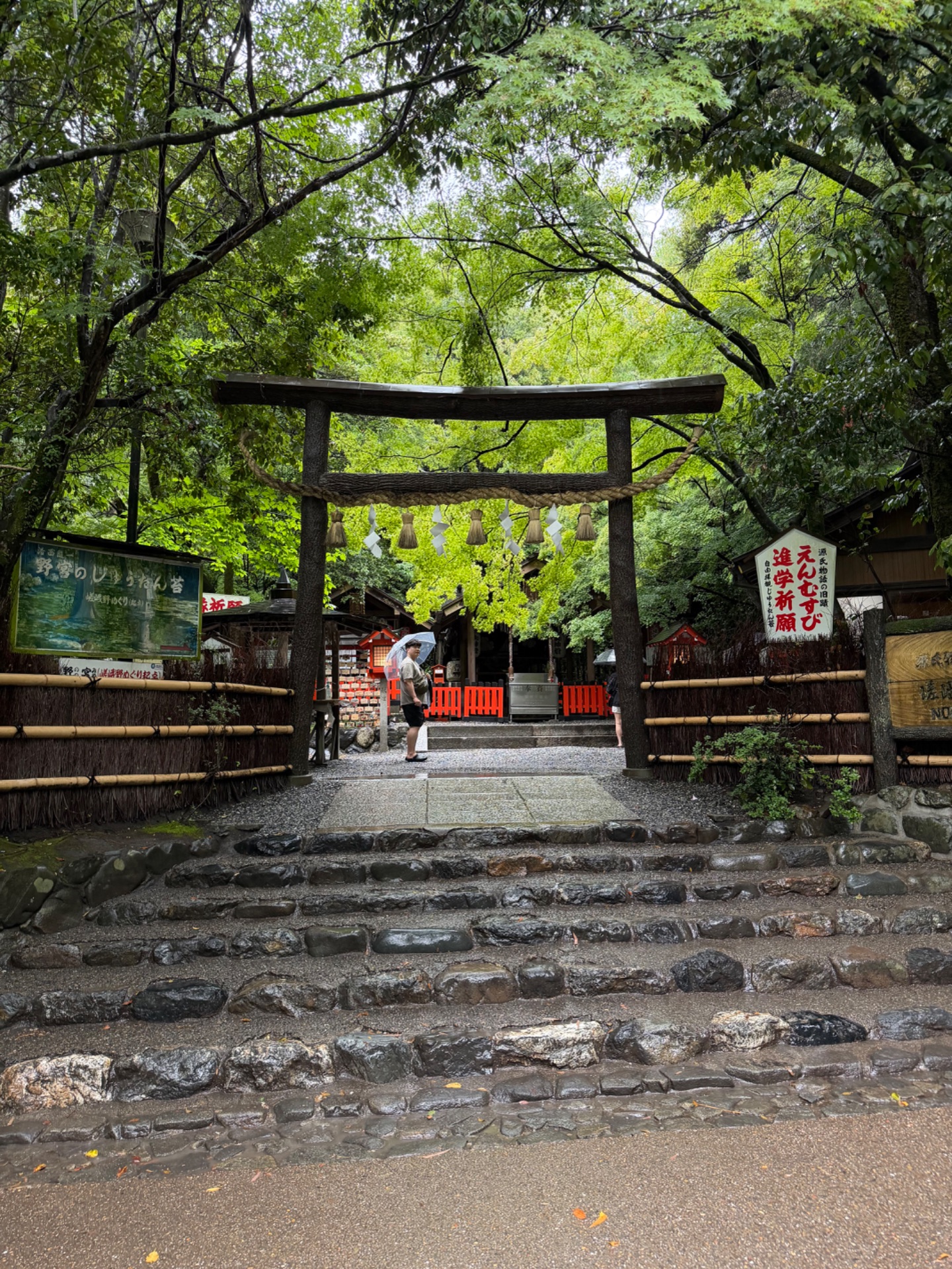 Japanese shrine entrance with wooden torii gate framed by lush green trees and stone steps leading to red lanterns.