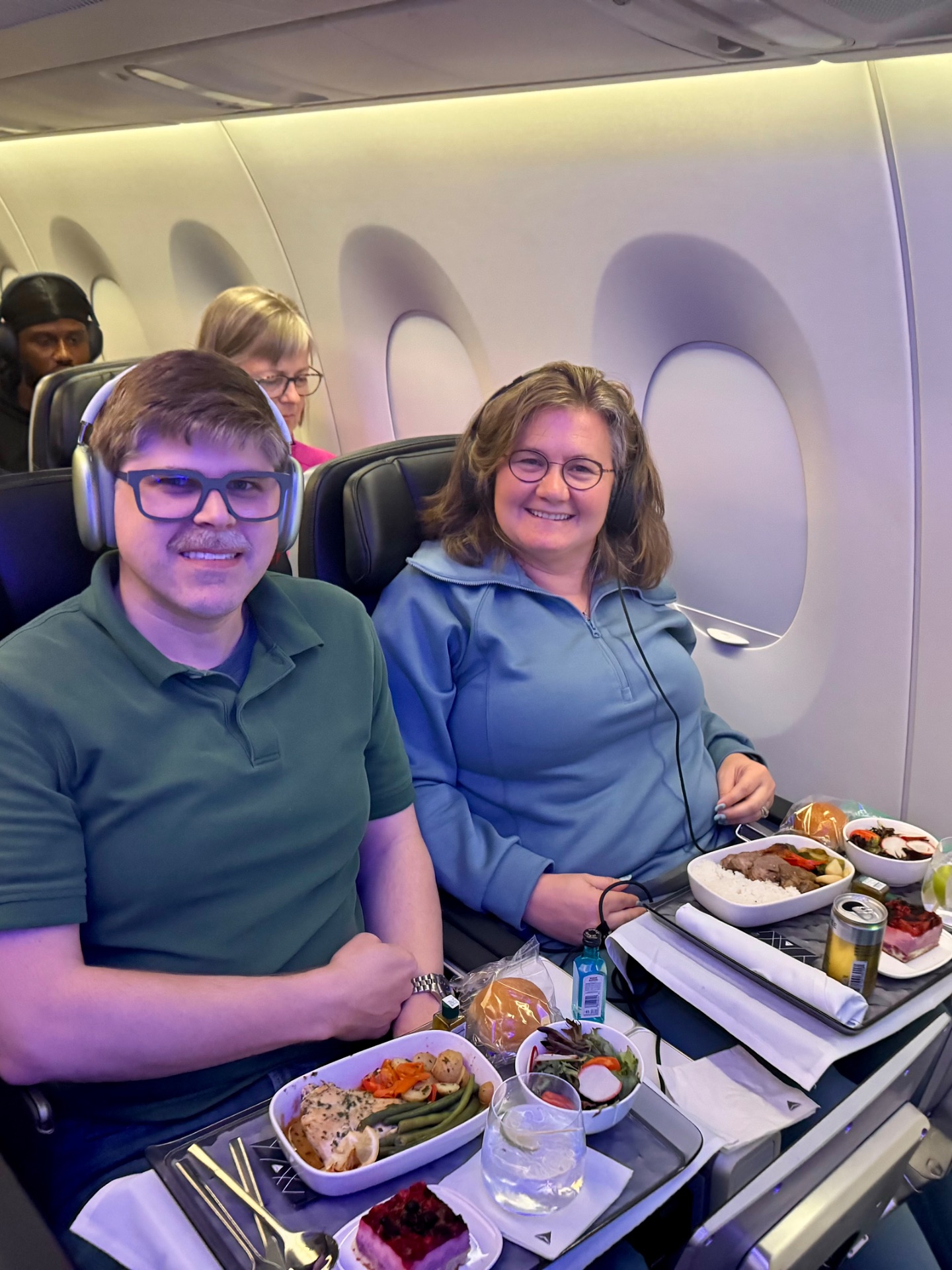 Two passengers enjoying meal service on an airplane with window seats visible in background.