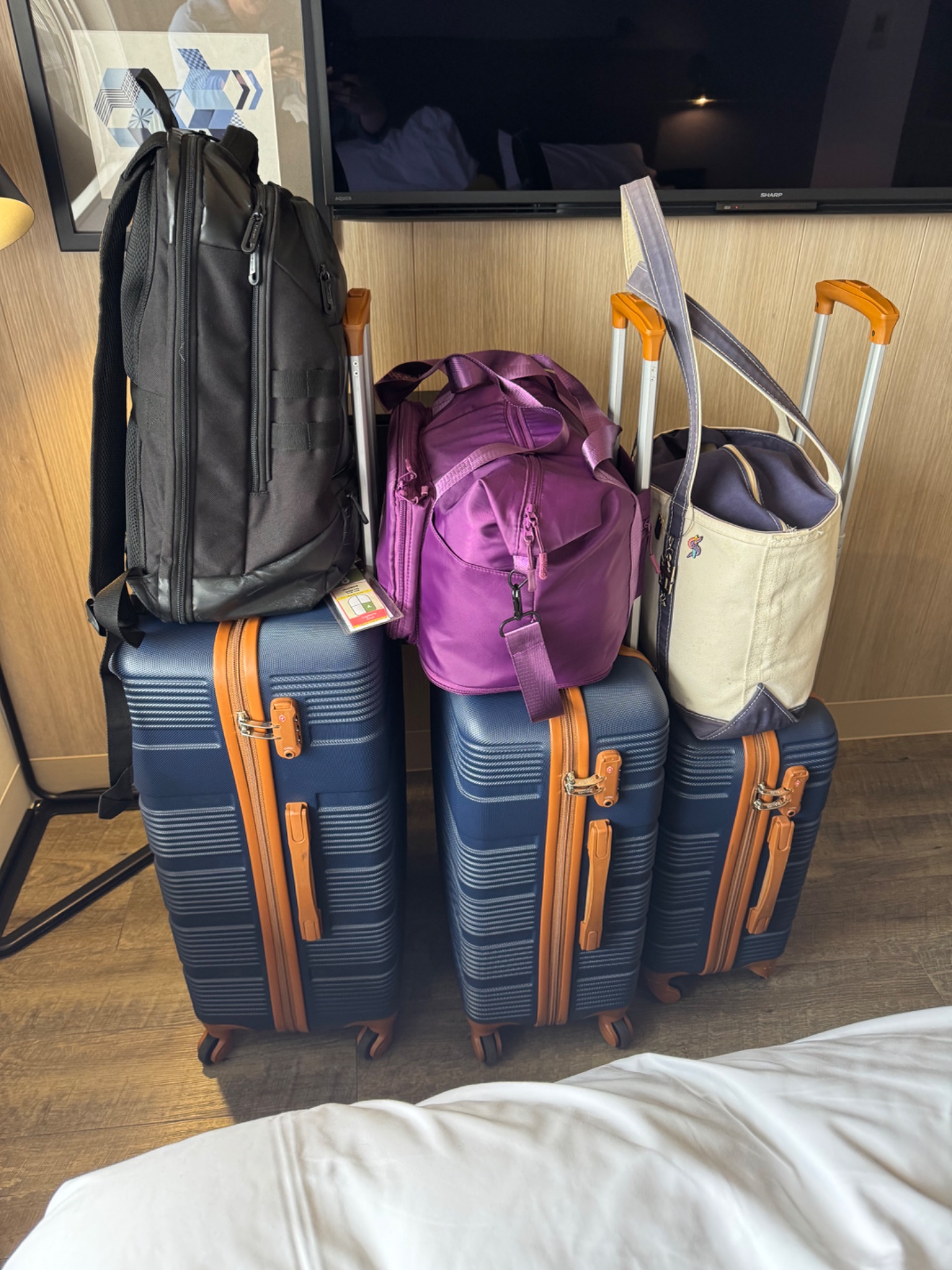 Display of luggage and travel bags at a cruise ship cabin, including three blue suitcases with orange accents, black backpack, purple backpack, and cream tote bag.