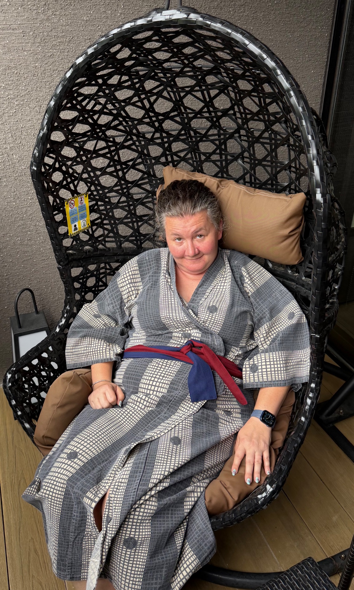 Woman in gray kimono sitting in black woven hanging egg chair with tan cushion indoors