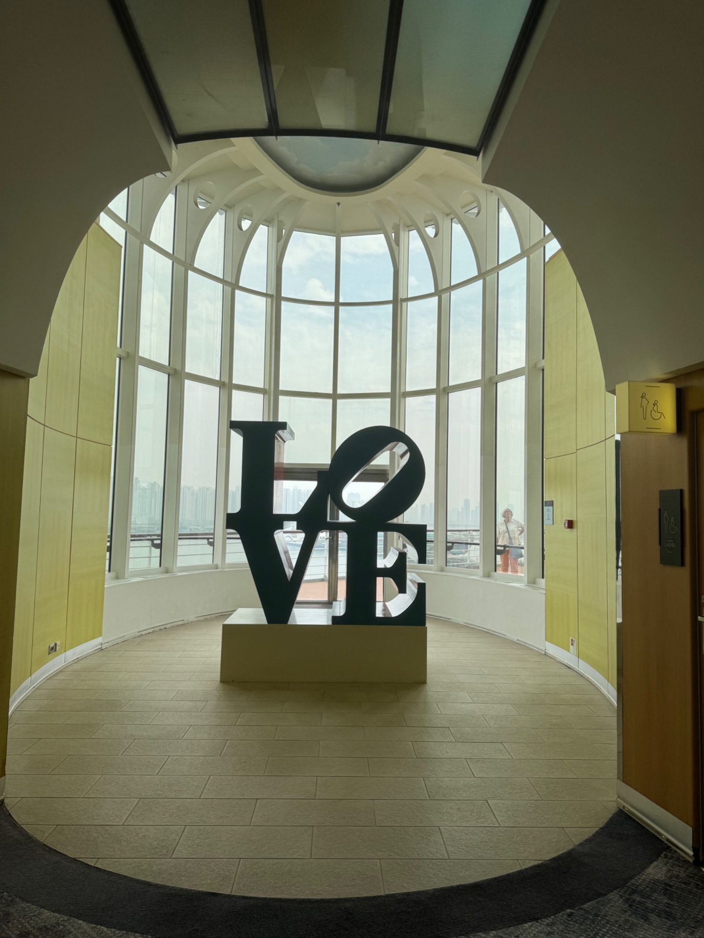 LOVE sculpture displayed in a modern gallery space with curved windows overlooking a cityscape and water view.