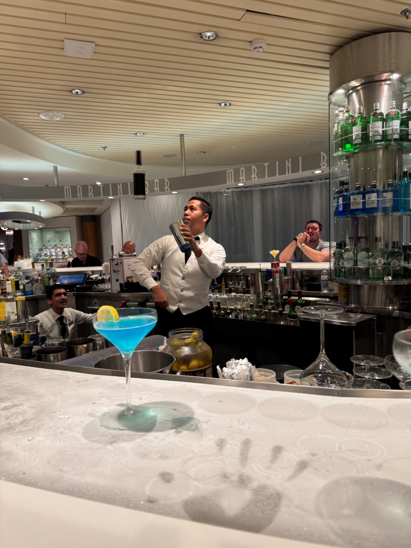 Bartender demonstrating cocktail mixing at elegant cruise ship martini bar with blue cocktail on marble counter.