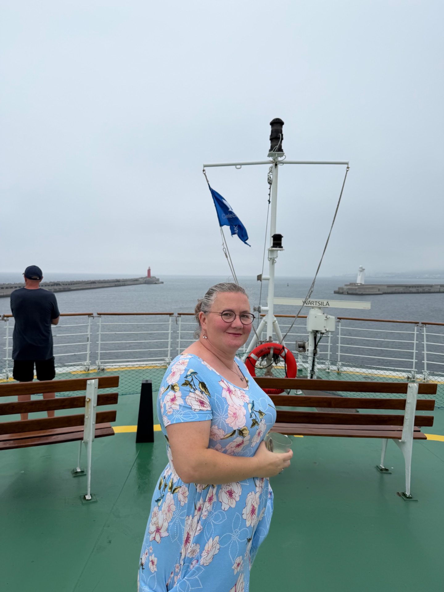 Woman on ferry deck with blue flag, harbor lighthouses and breakwater visible in background.