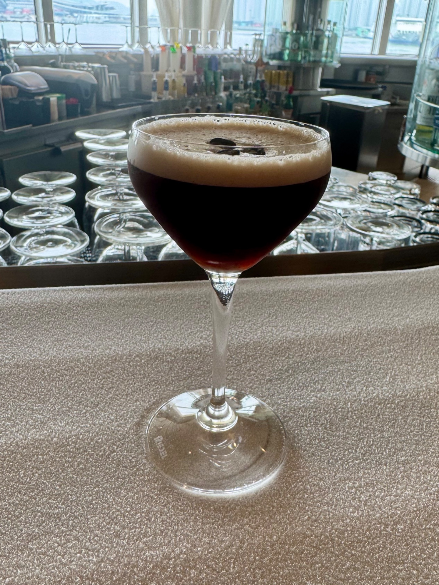 Dark cocktail with thick foam head in coupe glass on cruise ship bar counter with ocean view and bottles behind.