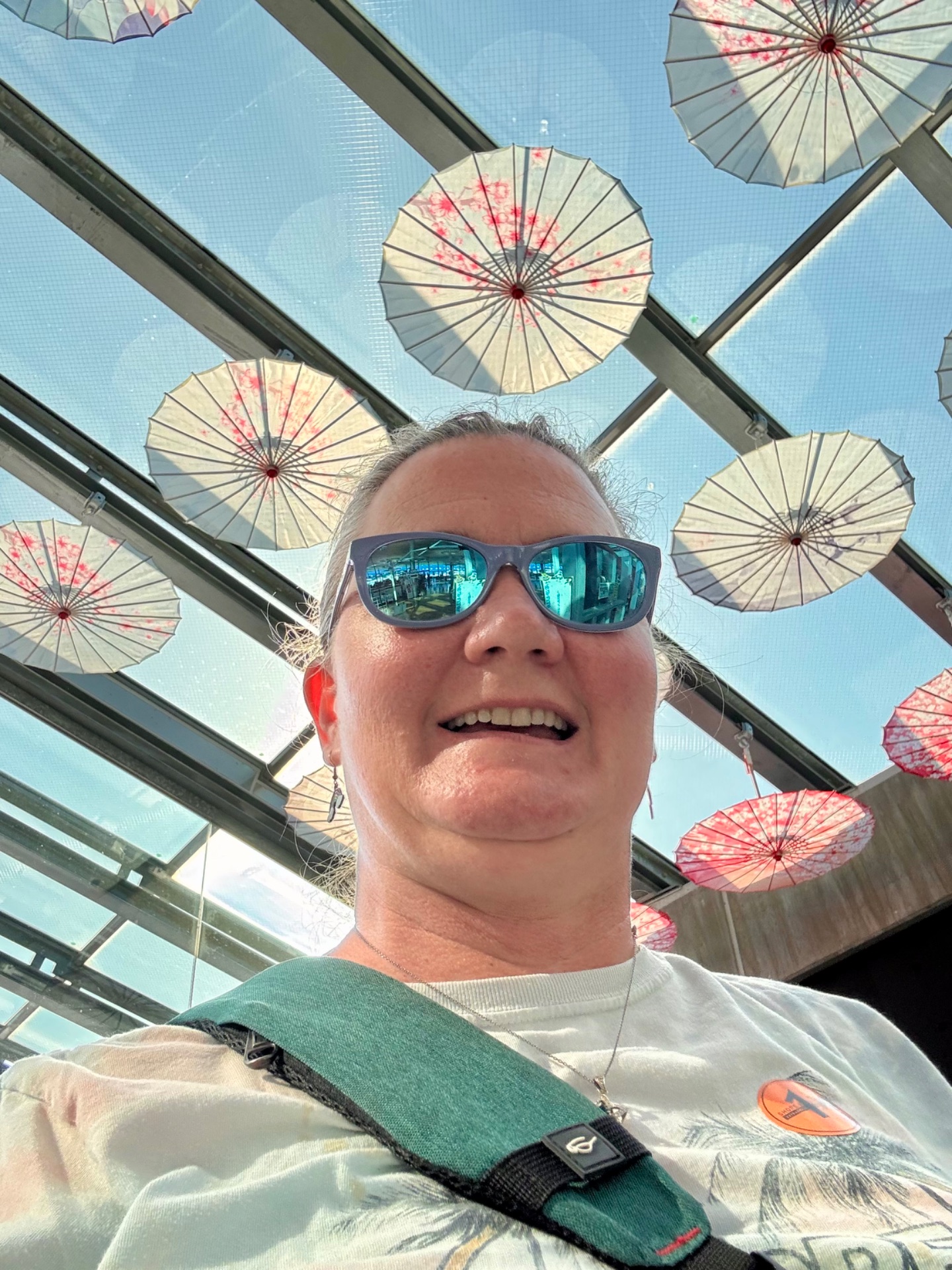 Man wearing sunglasses smiling under decorative umbrellas on a cruise ship deck with blue sky above.