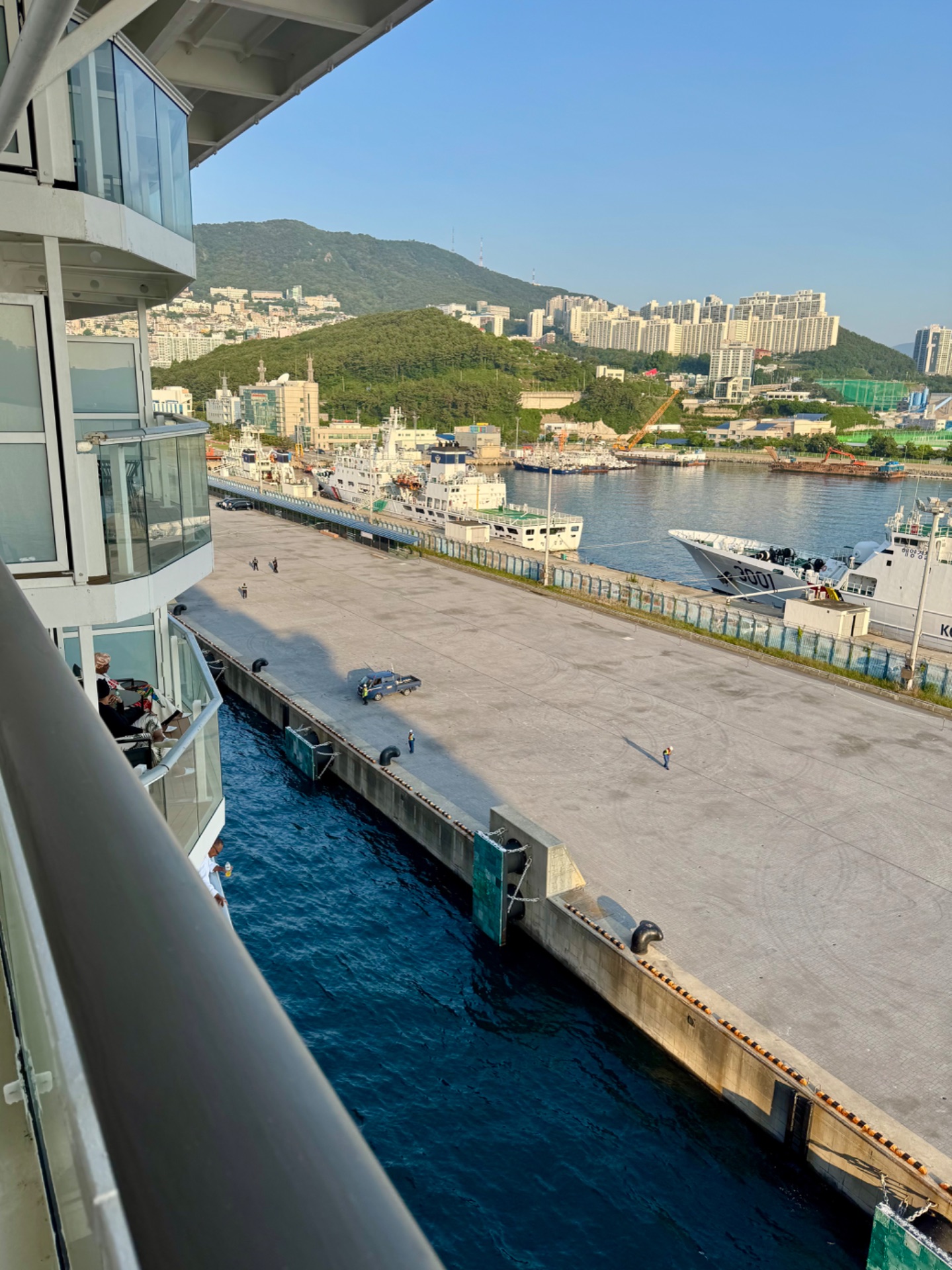View from cruise ship balcony of port with cargo vessels, mountainside city, and docked ships in harbor.