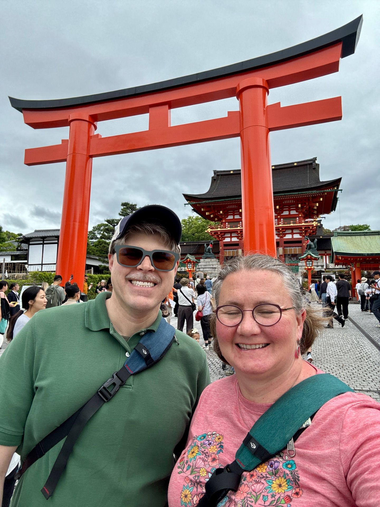 Two tourists smiling at a Japanese temple with a large red torii gate and traditional architecture in the background.