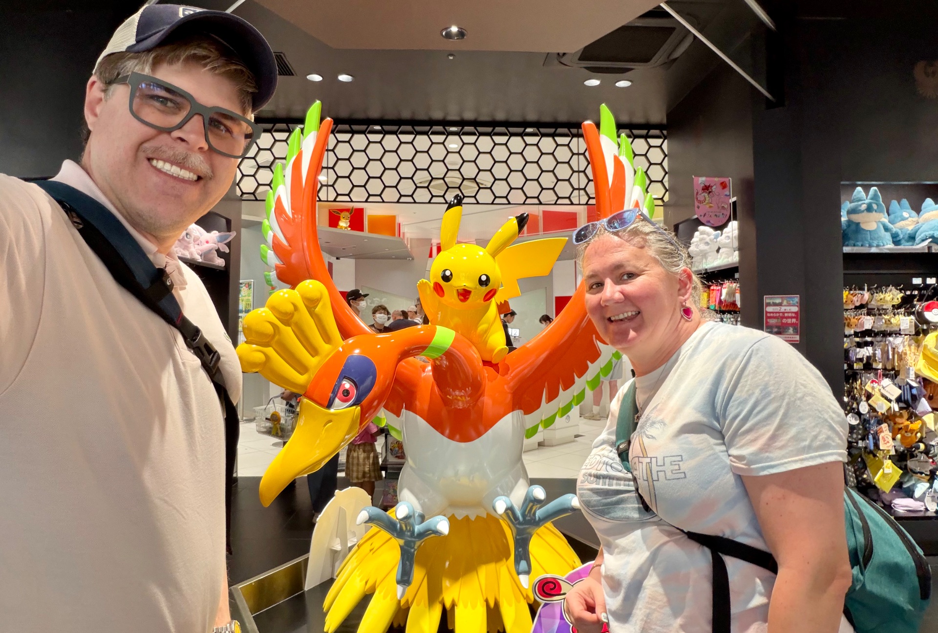 Two visitors posing with a large colorful Ho-Oh Pokémon statue in a Pokémon Center gift shop.
