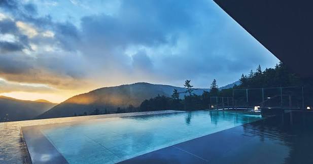 Infinity pool overlooking mountain lake at sunset with forested peaks and golden sky reflected in calm blue water