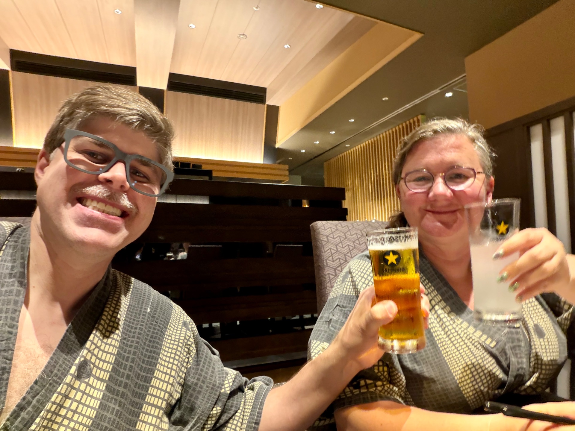 Two men wearing glasses toasting with beer in an elegant cruise ship lounge with warm lighting and piano in background