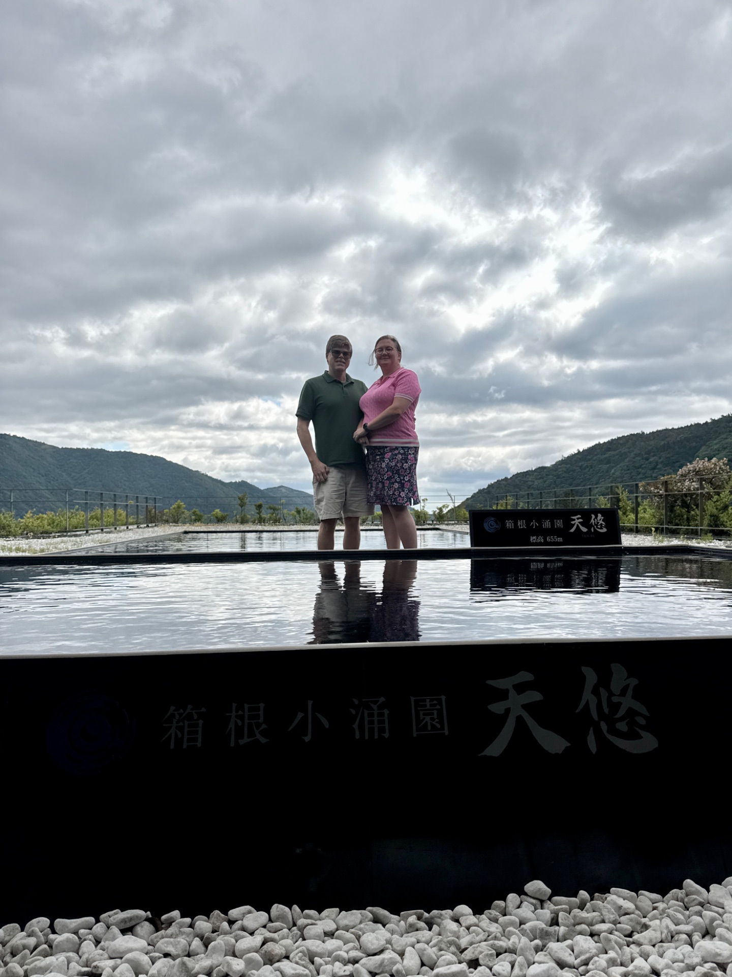 Two tourists pose at a scenic riverside overlook with mountains, water features, and Chinese characters visible in the landscape.