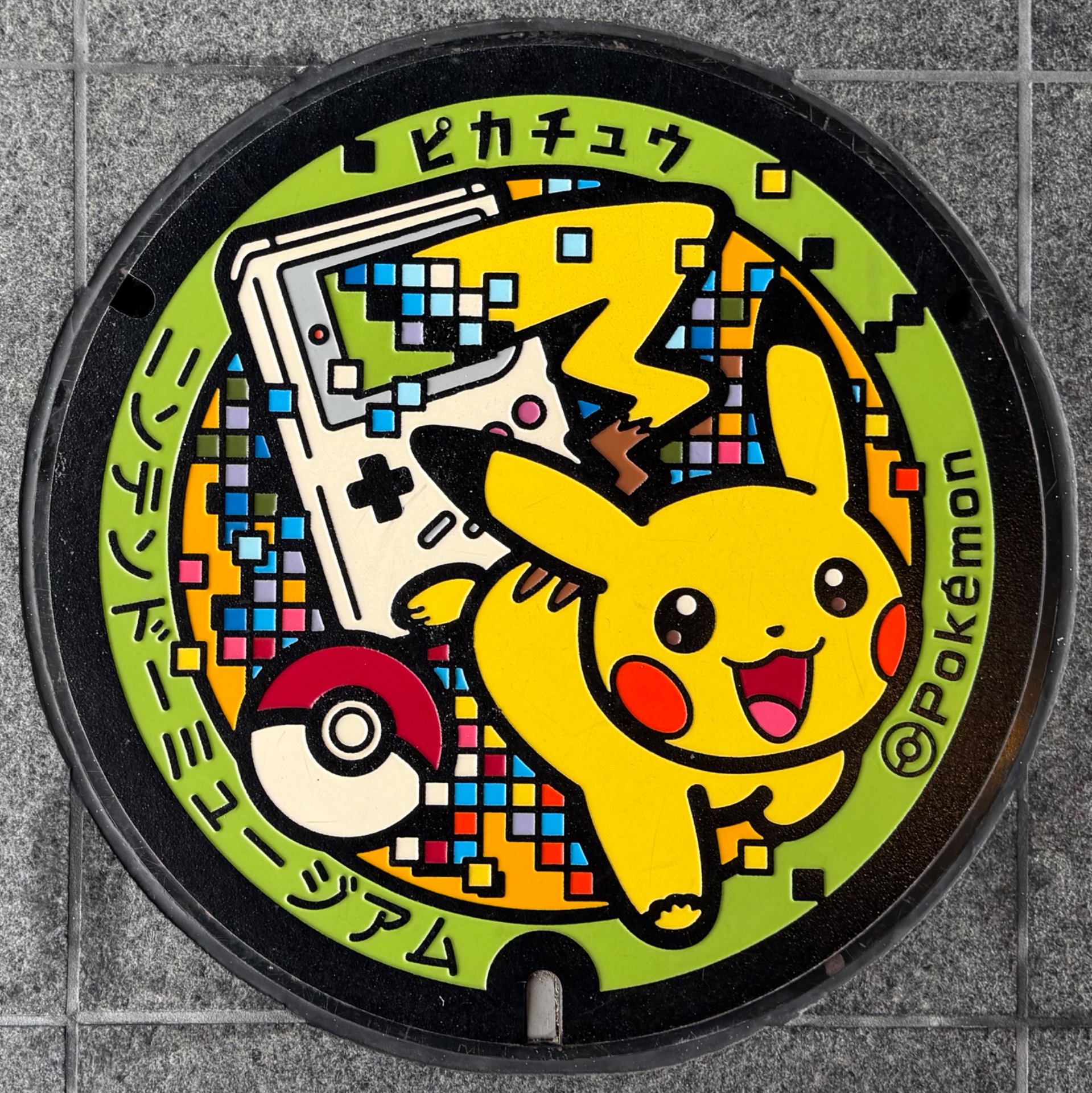 Pokémon-themed manhole cover featuring Pikachu with a Game Boy and Poké Ball on green background in Japan.