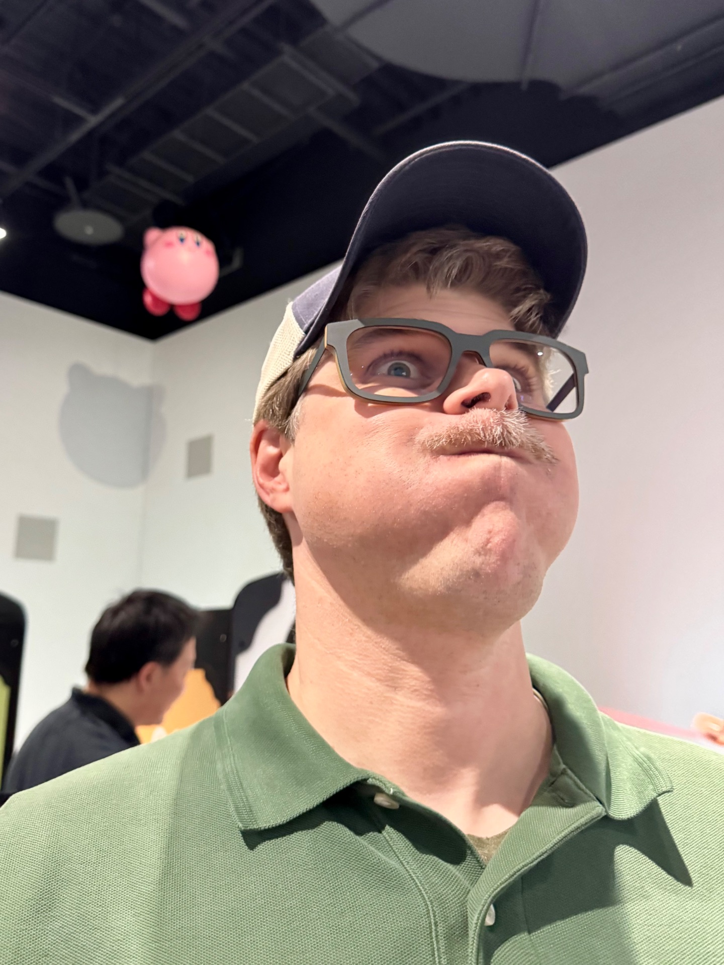 Man wearing glasses and navy cap in green shirt looking upward at pink decorative object in indoor venue