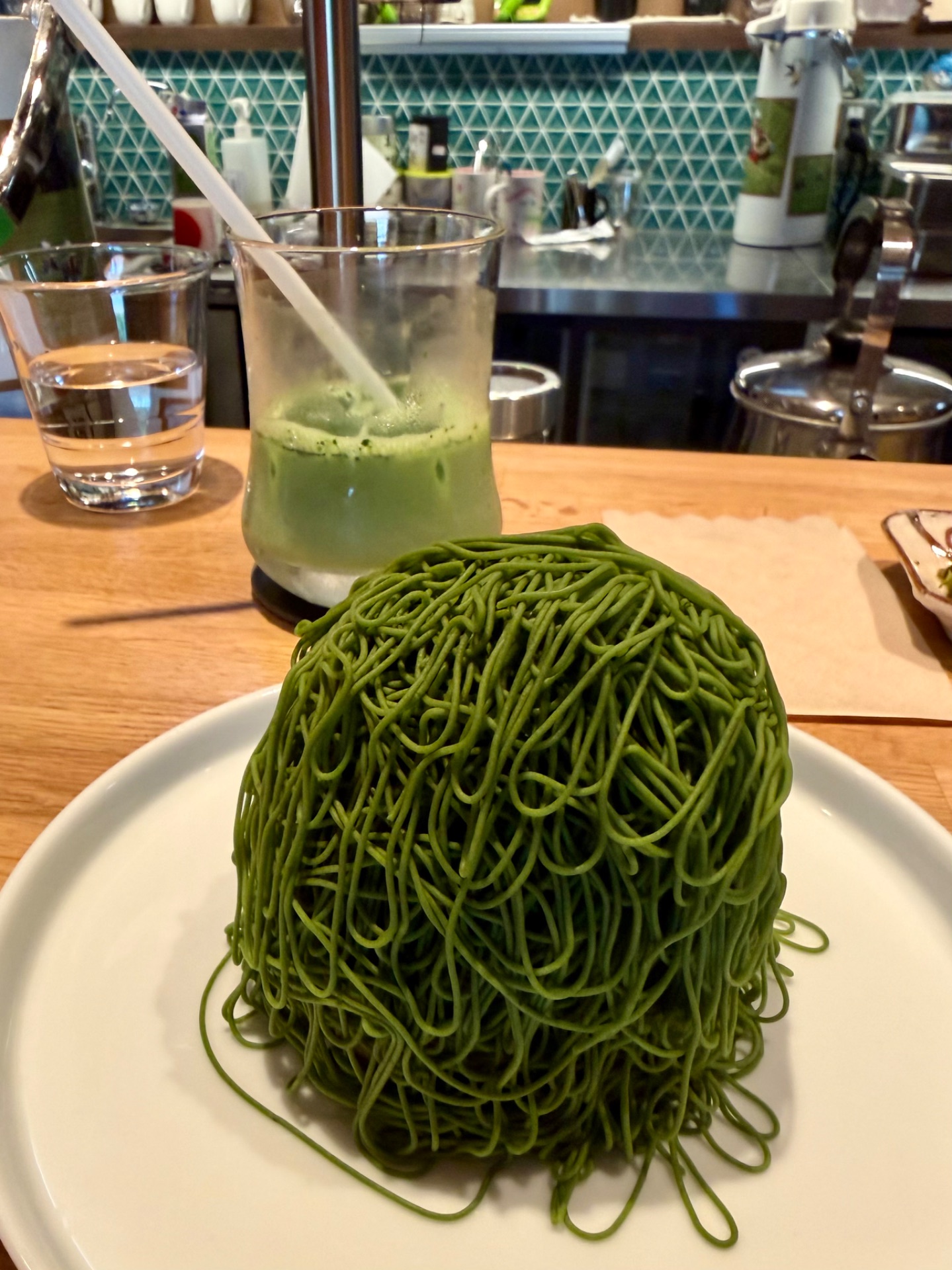 Vibrant green noodle dish served on white plate at upscale restaurant with matcha beverage in background.