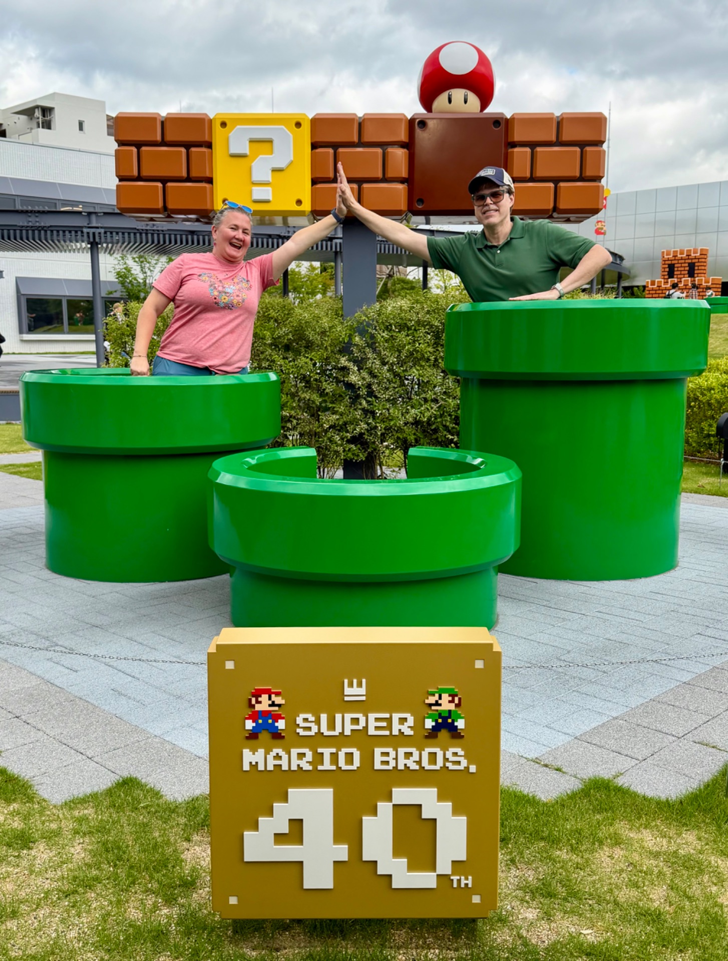 Two visitors pose at a Super Mario Bros. 40th anniversary installation with green pipes and iconic game elements.
