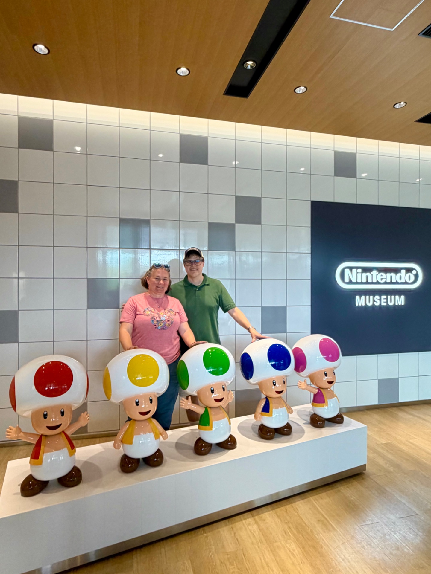 Two visitors pose behind colorful Toad character figures at the Nintendo Museum with illuminated logo sign.