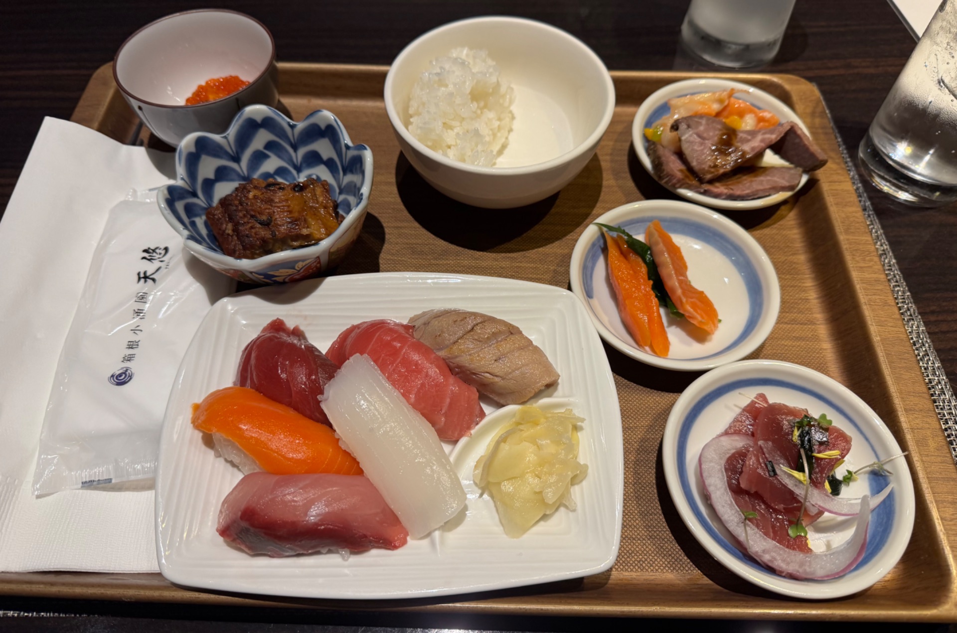 Japanese sushi and sashimi platter with rice, pickled vegetables, and side dishes served on a wooden tray.