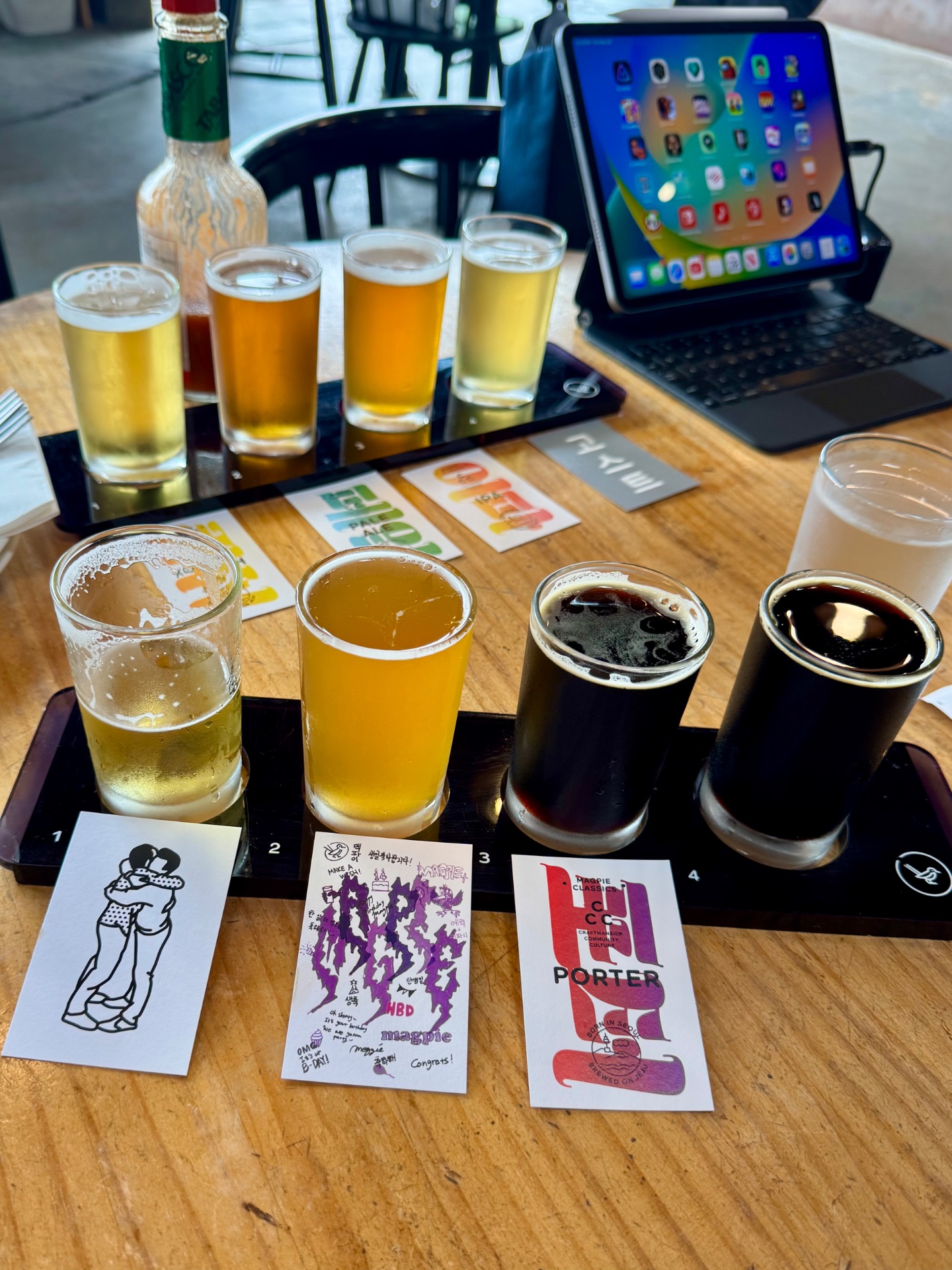 Beer flight tasting at a brewery with four different samples labeled 1-4, ranging from light to dark, displayed on a wooden table with an iPad nearby.