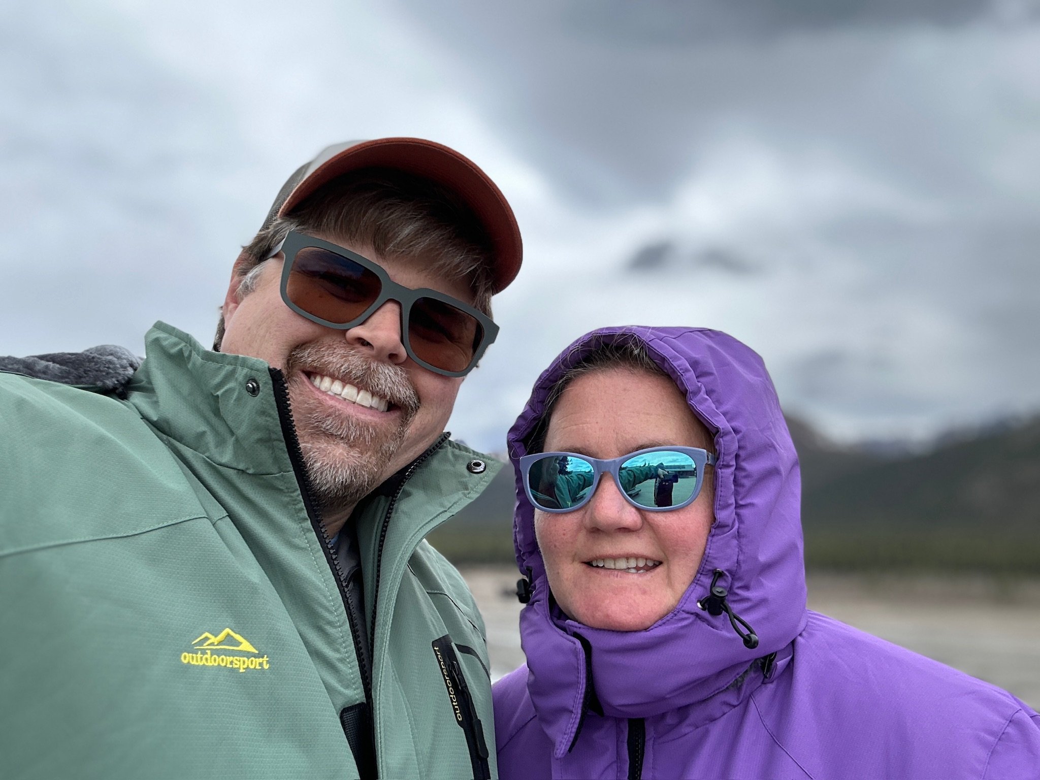 Two people smiling outdoors wearing sunglasses and rain jackets