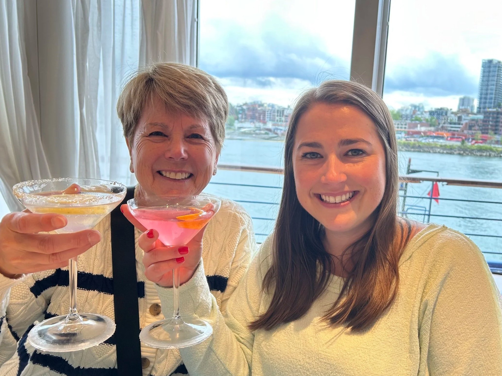 Two women toasting cocktails on cruise ship balcony overlooking waterfront city