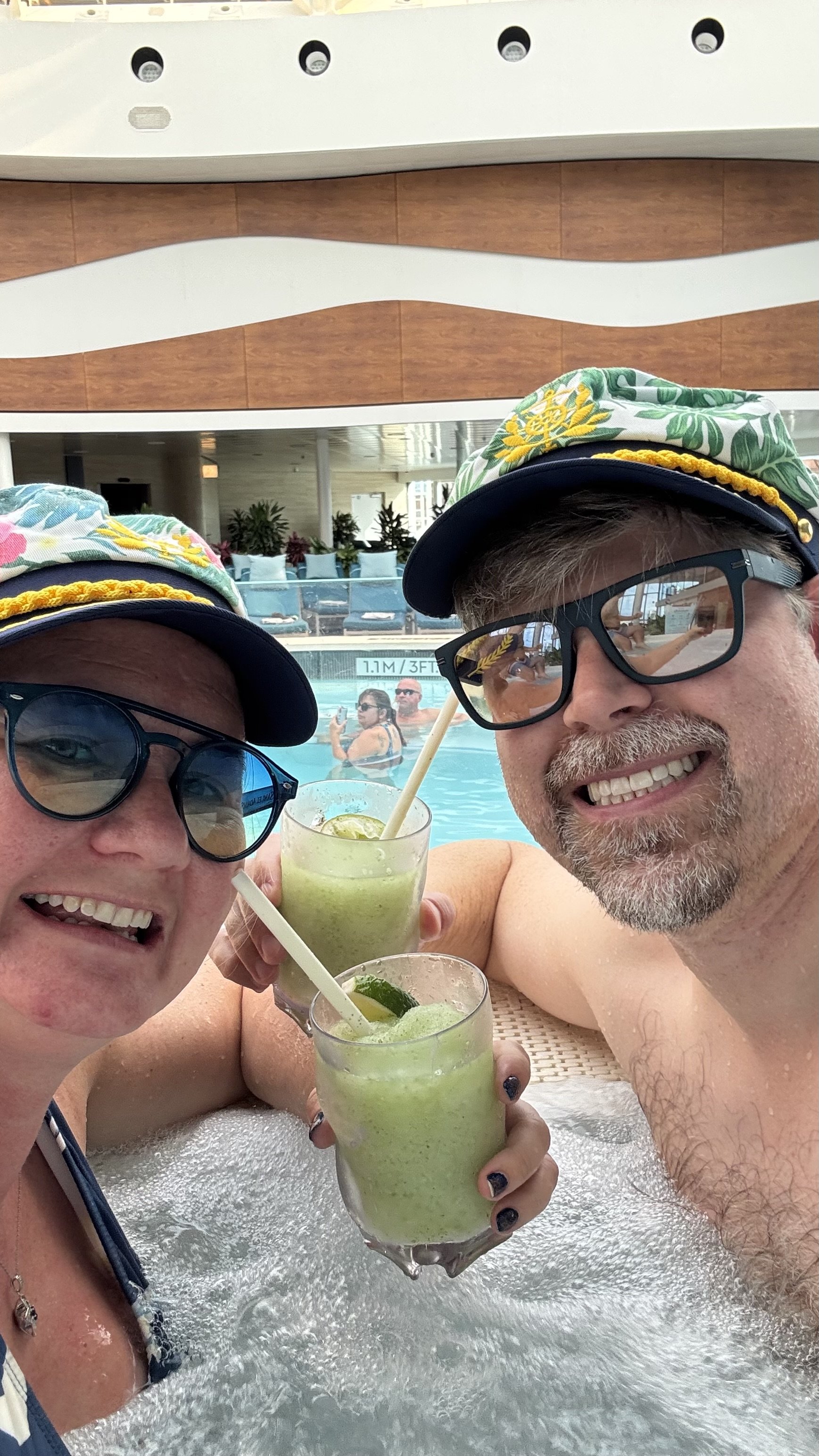 Two people in tropical hats and sunglasses holding mojitos by a cruise ship pool deck.