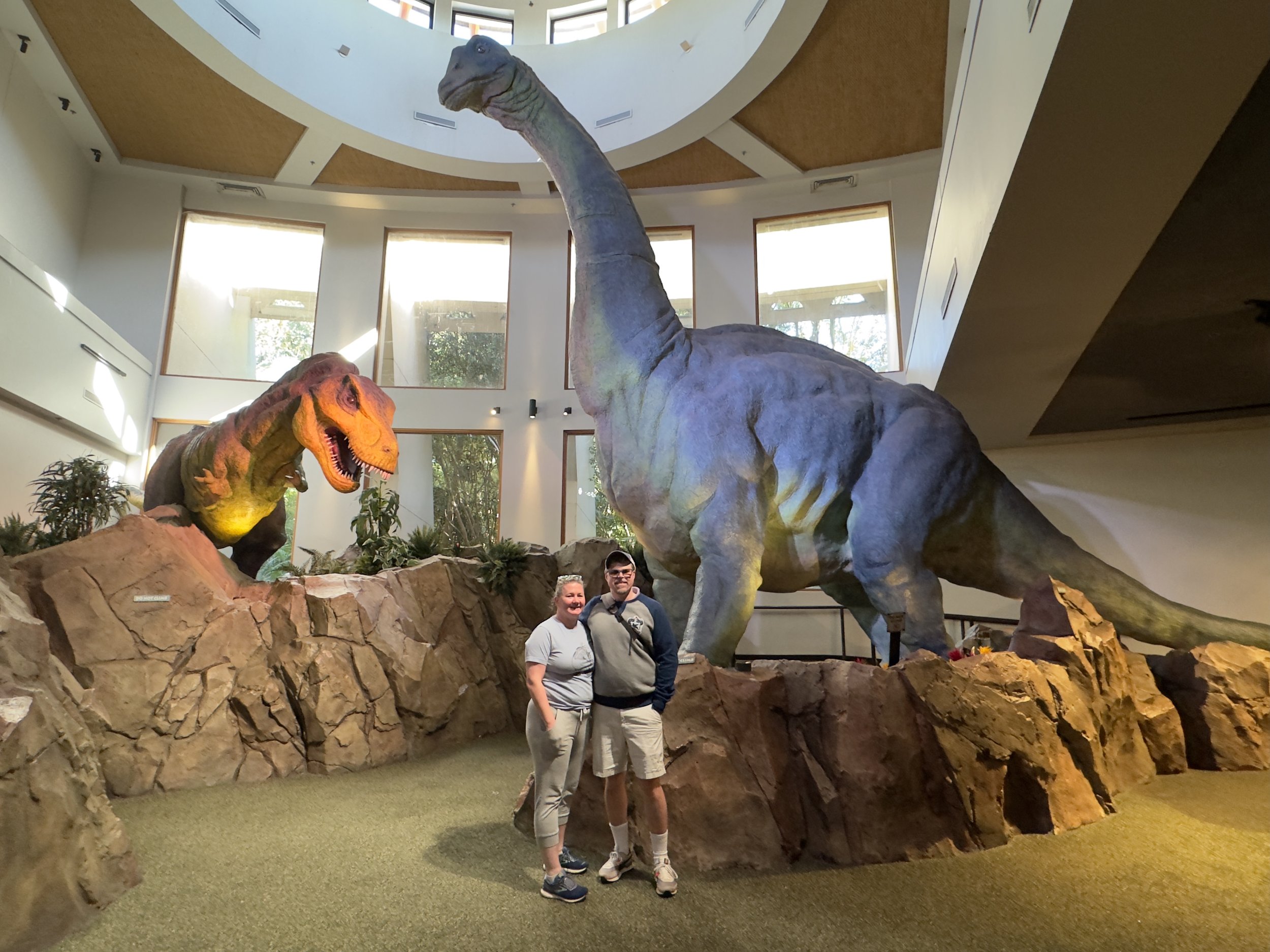 Two visitors pose with life-size dinosaur exhibits in museum atrium